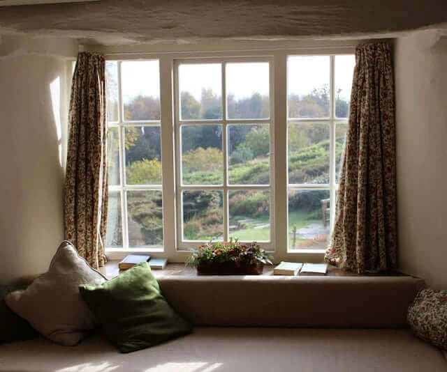 Cozy window view with floral curtains, cushions, and greenery outside, creating a serene atmosphere for relaxation.