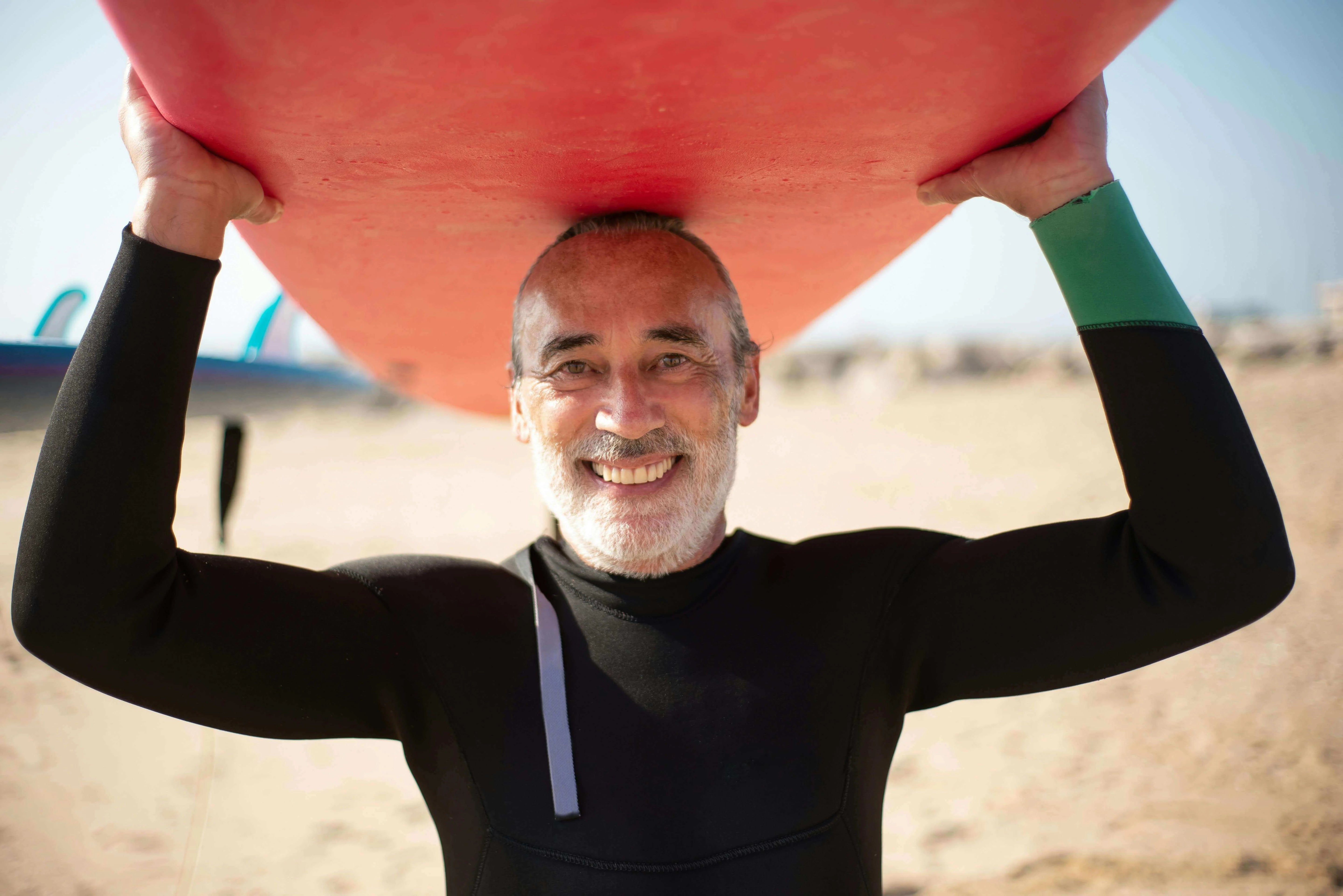 Happy man in wetsuit holding a red surfboard over his head on the beach, enjoying life and water sports.