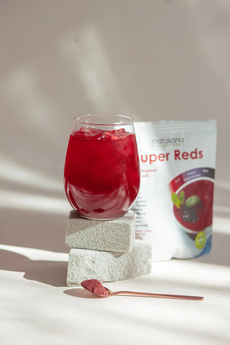 Matakana Superfoods Organic SuperReds 120g in a glass with ice beside a packet and spoon on a textured background.