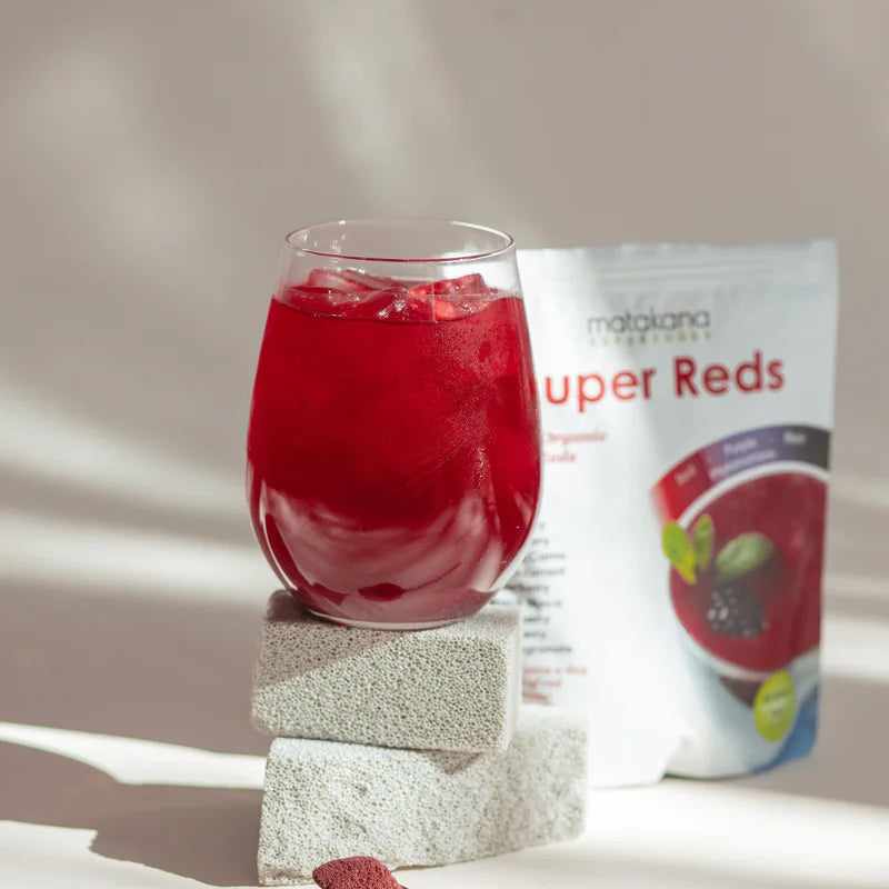 Matakana Superfoods Organic SuperReds 120g in a glass with ice beside a packet and spoon on a textured background.