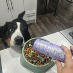Dog looking at a bowl of food with a person holding a 'Fourflax' container in a kitchen.